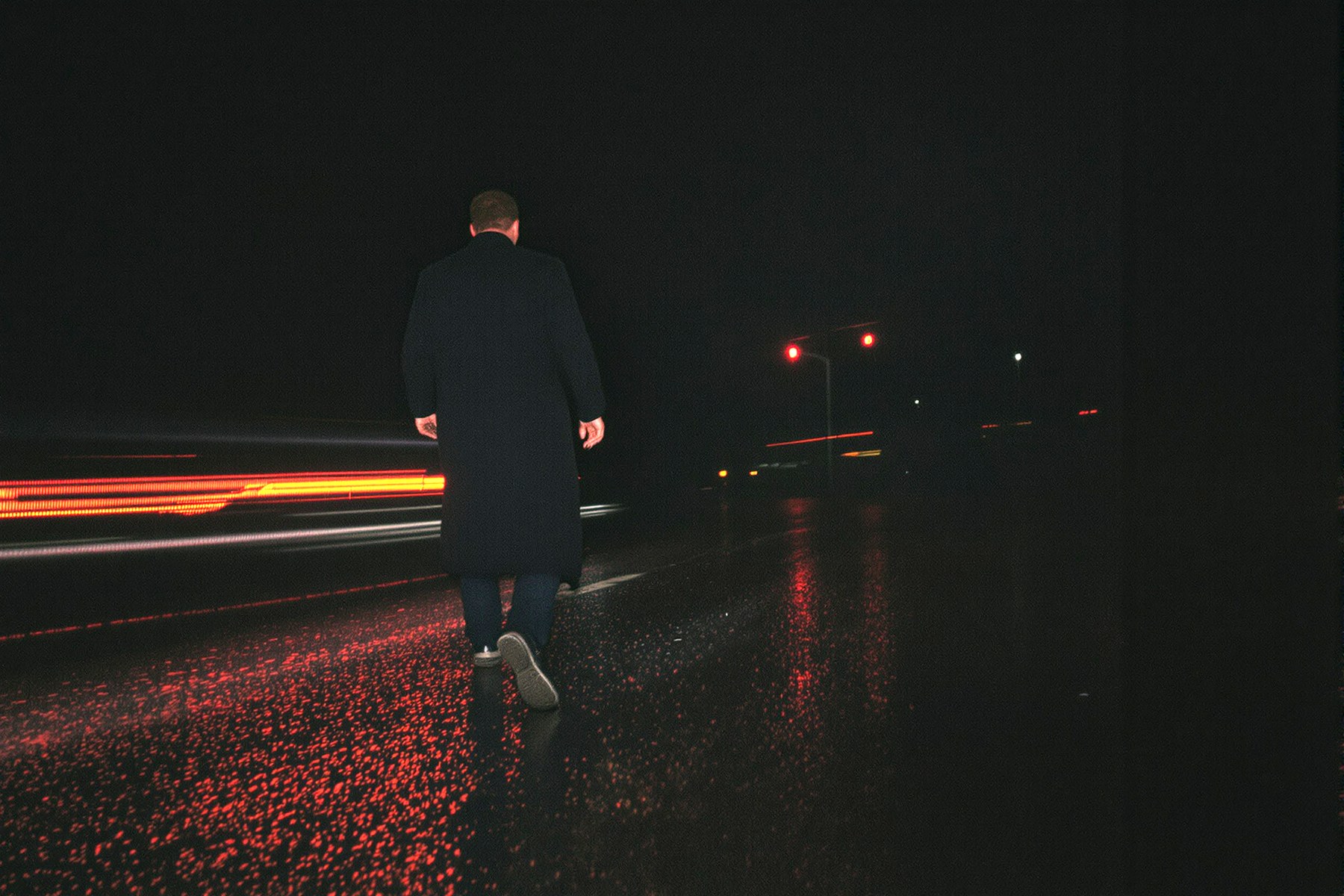A man in a long dark coat walking across wet asphalt at night, red tail lights streaking past.
