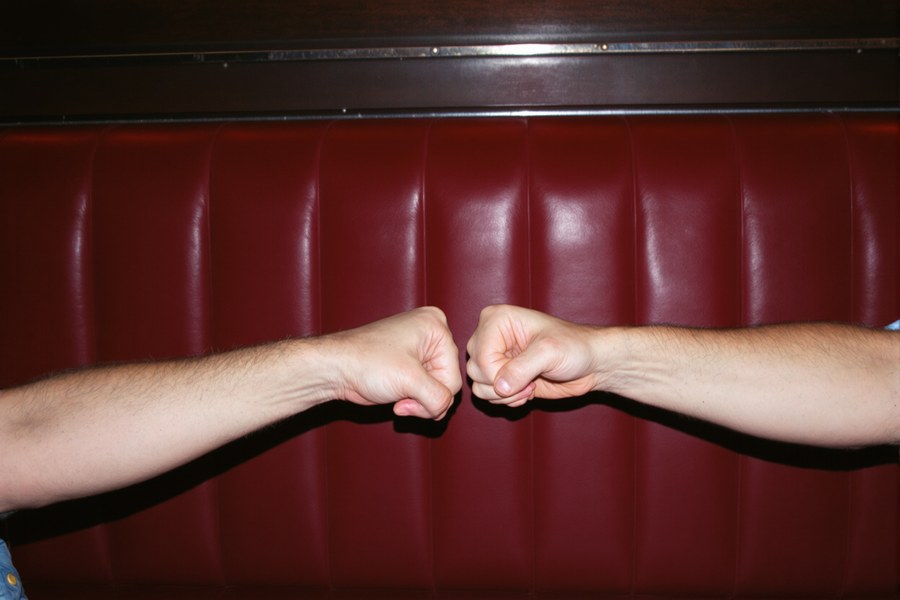 Two people bumping fists across a dark red leather booth at night.