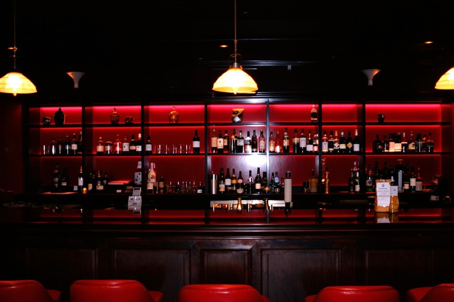 An empty bar at night, red stools along a dark counter under one warm lamp.